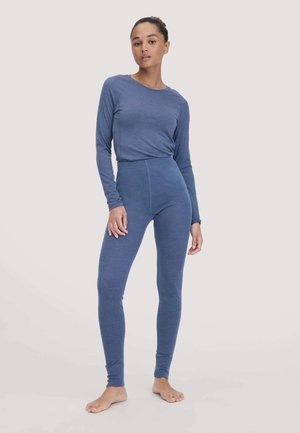 Eine Frau steht barfuß und trägt ein passendes blaues Langarmoberteil und eng anliegende Leggings vor einem hellen Hintergrund.