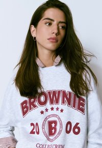 Sudadera gris con "BROWNIE" en grandes letras rojas, con un logo redondo y estrellas. El cuello a rayas añade textura. Modelo con cabello largo.