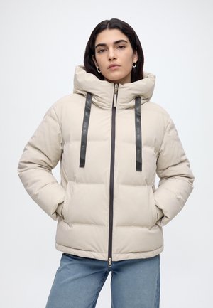 Donna con capelli scuri che indossa una giacca imbottita beige con colletto ampio e cordini neri, mani nelle tasche, in piedi su sfondo bianco.