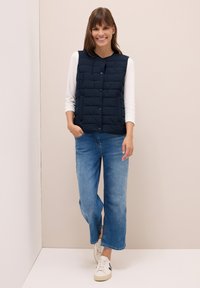 Gilet imbottito blu navy con motivo trapuntato, con bottoni a scatto sul davanti, indossato sopra una maglietta bianca a maniche lunghe, abbinato a jeans azzurri chiari e scarpe da ginnastica bianche.
