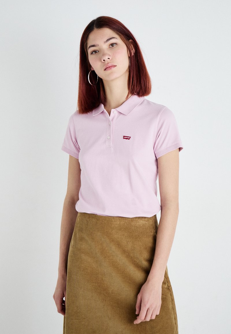 Camisa polo rosa con cuello y mangas cortas, que presenta un pequeño logo rojo de Levi's en el pecho, combinada con una falda de pana marrón.