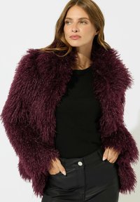 Giacca di pelliccia sintetica bordeaux con una texture shaggy, dotata di un ampio colletto e maniche lunghe, abbinata a una maglietta nera e pantaloni neri.