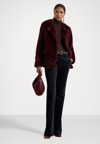 Burgundi színű műszőrme kabát, kivágott gallérral, barna garbó, sötétkék kordbársony nadrág és egy passzoló kézitáska, amely sima textúrát mutat.