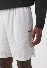 Hvide mesh sportsshorts med et sort Nike-logo, der har en tekstureret overflade og vandrette sømdetaljer nær kanten.