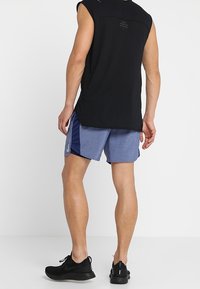 Svart ärmlös atletisk topp med mesh-textur, tillsammans med blå och marinblå shorts med en logotyp. Bärs med svarta sportskor.