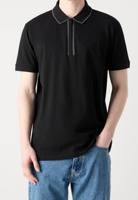 Homme portant un polo noir à manches courtes avec un détail de fermeture éclair et un jean bleu, une main dans la poche, debout devant un fond blanc.