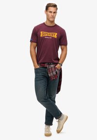 Bordeaux t-shirt met "SUPERDRY SUPPLY CO." graphic, blauwe logo patch, gecombineerd met vervaagde blauwe jeans en beige sneakers, met een geruite overhemd om de taille geknoopt.