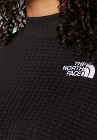 Svart texturerad tröja med hög krage och ett upphöjt rutmönster; har en broderad vit logotyp på vänster bröst.