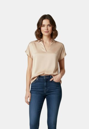 Žena s hnedými vlasmi po ramená, oblečená v béžovej krátkorukávovej blúzke a tmavomodrých rifle vysokého pásu, stojaca s jednou rukou v vrecku.