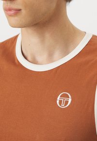 Jeune personne portant un débardeur couleur rouille avec un bord blanc et un petit logo blanc circulaire sur la poitrine.