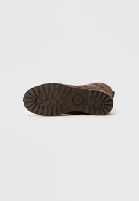 Suela de bota marrón con surcos profundos para tracción, material de goma, y un logo circular. Superficie lisa con una textura ligeramente elevada.