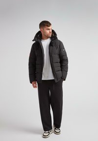 Mand iført en sort polstret jakke over en hvid T-shirt, sorte bukser og sort-hvide sneakers, der står foran en lysegrå baggrund.