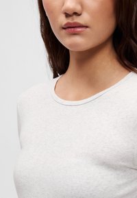 Šviesiai pilka ilgomis rankovėmis marškinėliai su apvaliu kaklu. Audinys atrodo minkštas, su šiek tiek tekstūruota paviršiaus struktūra ir subtiliu margu raštu.