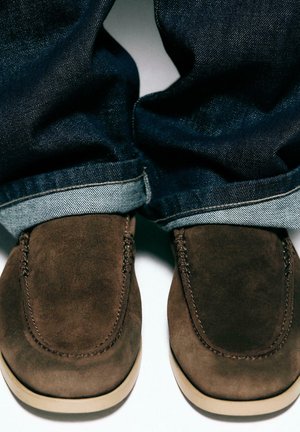 Chaussures marron en daim à enfiler portées avec un jean bleu foncé retroussé sur une surface blanche.