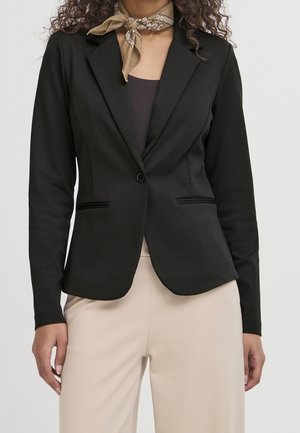 Femme portant un blazer noir cintré, un pantalon beige, un haut foncé et un foulard beige à motifs autour du cou, debout devant un fond clair uni.