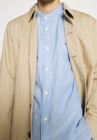 Homme portant une chemise boutonnée bleu clair sous un trench-coat beige, se tenant devant un fond blanc uni.