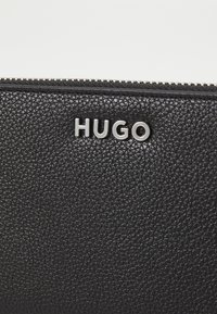 Zwart leren portemonnee met textuur, voorzien van een ritssluiting en zilveren "HUGO" merknaam aan de voorkant. Glad hardware accent.