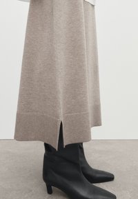Moitié inférieure d'une jupe en maille beige avec une fente latérale portée sur des bottes noires à talons moyens et bouts pointus sur un sol gris.