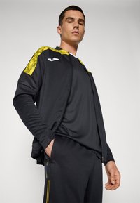 Zwarte sportjack met gele geprinte schouderaccenten. Het model draagt een bijpassende zwarte top en broek, waarmee een gestroomlijnde pasvorm wordt getoond.