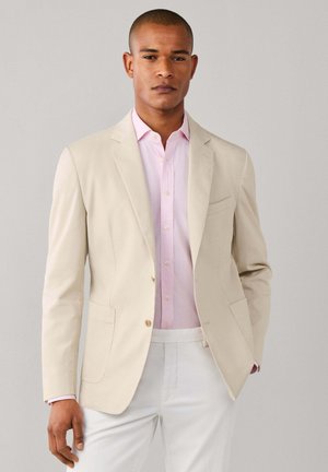 Man draagt een beige blazer, lichtroze overhemd en witte broek, staand tegen een effen grijze achtergrond.