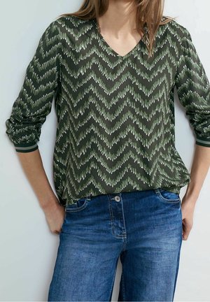 Groene blouse met een V-hals en lange mouwen, voorzien van een zigzagpatroon in lichtere groentinten, gecombineerd met blauwe denim jeans.