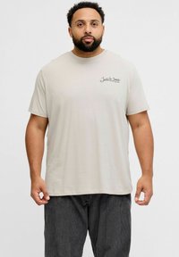 Jasnobeżowa koszulka z krótkim rękawem, z okrągłym dekoltem i małym czarnym logo na lewej piersi. Wykonana z miękkiej bawełnianej tkaniny.