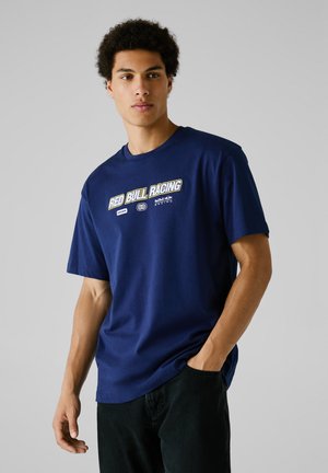 Jonge man met krullend haar die een marineblauw "Red Bull Racing" t-shirt en zwarte broeken draagt, met één hand in zijn zak, tegen een effen achtergrond.
