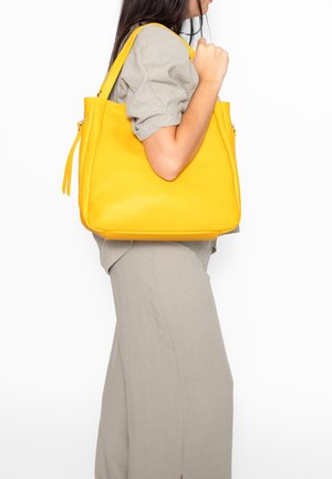 Donna con indosso un completo beige chiaro che porta una grande borsa tote gialla in pelle sulla spalla su sfondo bianco.