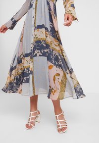 Robe à motif floral avec des manches longues, un bouton-pression à l'avant, et une jupe fluide dans des nuances de bleu, rose et vert. Sandales blanches à talons.
