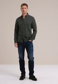 Camisa de botón de color verde oscuro, estilo ajustado, combinada con jeans azules y zapatos negros. Diseño simple, tela suave y detalles sutiles.