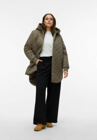 Veste longue matelassée vert olive avec un col montant, associée à un haut blanc et un pantalon large noir. Des chaussures jaunes ajoutent un contraste de couleur.