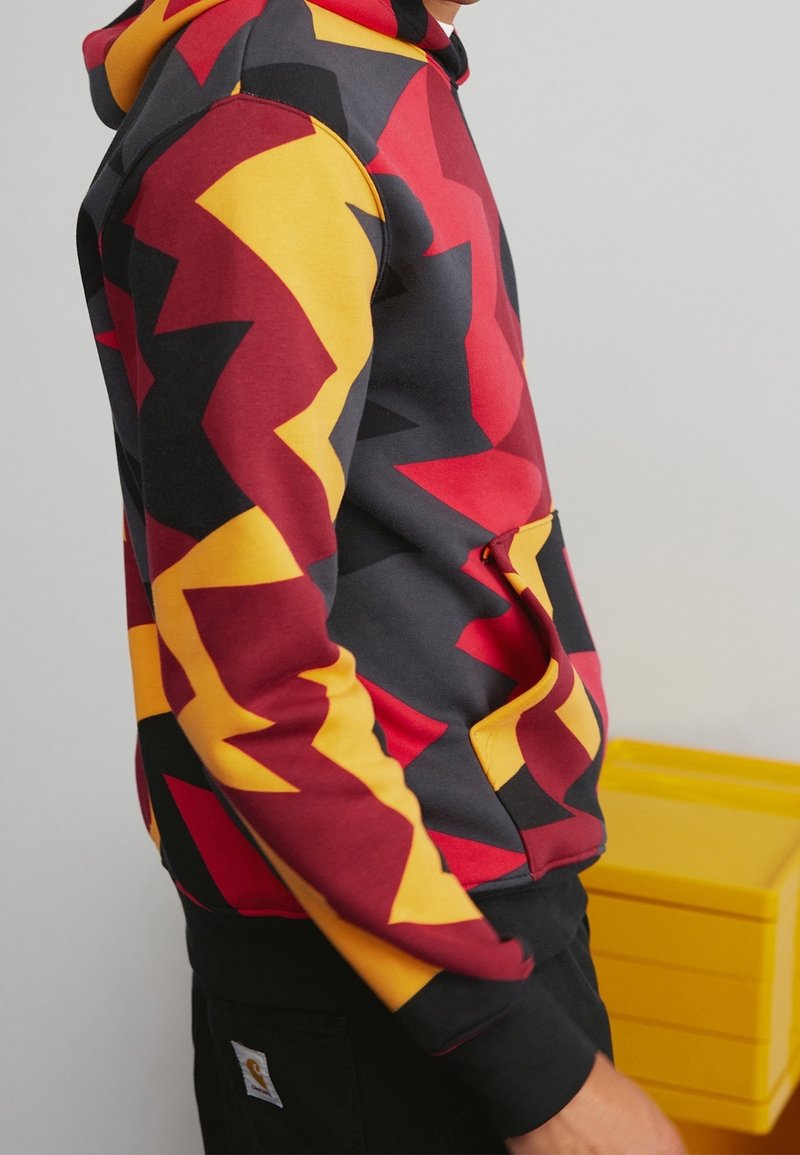 Sweat à capuche coloré avec un motif en zigzag audacieux en rouge, jaune et noir. Comprend une poche avant et des poignets côtelés.