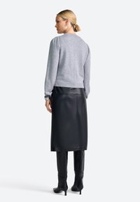 Femme aux cheveux blonds en chignon bas portant un pull gris, une jupe midi en cuir noir, des bottes noires jusqu'aux genoux, ainsi que des boucles d'oreilles et des bracelets en argent.