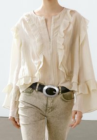 Light beige blouse with ruffled details and a zipper at the front, paired with light gray jeans and a black belt featuring a circular silver buckle.