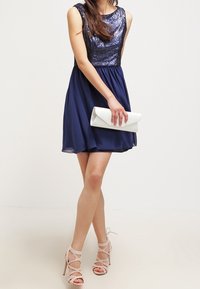 Námořnické šaty s flitrovou korzetovou částí, volnou sukní a bezrukávovým designem. Doplňuje je bílá kabelka a nahé sandály na remínkách.