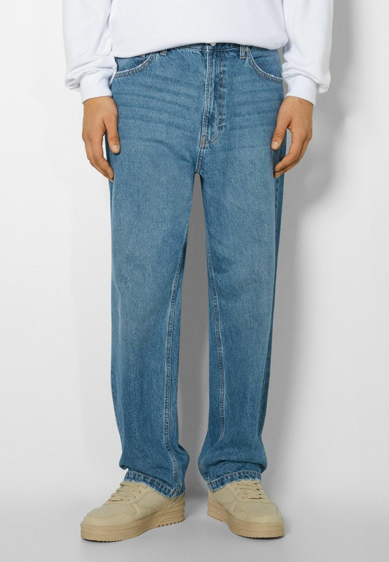 Jeans de mezclilla de color azul claro con un corte relajado, que presentan un diseño tradicional de cinco bolsillos y un ligero desvanecido, combinados con zapatillas de color claro.