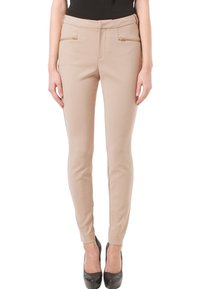 Nainen, joka on pukeutunut beigen värisiin slim-fit-housuihin, joissa on kultaiset vetoketjutaskut, mustaan toppiin ja mustiin korkokenkiin, seisoo valkoista taustaa vasten.