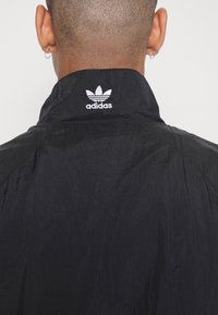 Svart lättviktsjacka med texturerad yta, med en broderad vit Adidas-logotyp på kragen. Minimalistisk design utan ytterligare detaljer.
