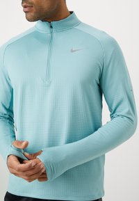 Jasnoniebieska, długorękawowa bluza z zamkiem, wykonana z fakturowanej tkaniny. Posiada otwory na kciuki w rękawach oraz małe szare logo Nike na piersi.