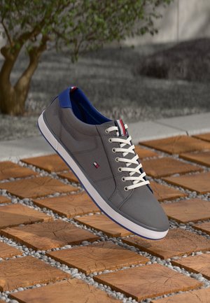 Grå casual sneaker med hvide snørebånd og blå indvendig foring, hviler på en træflise og en grå småstensbelægning udendørs.