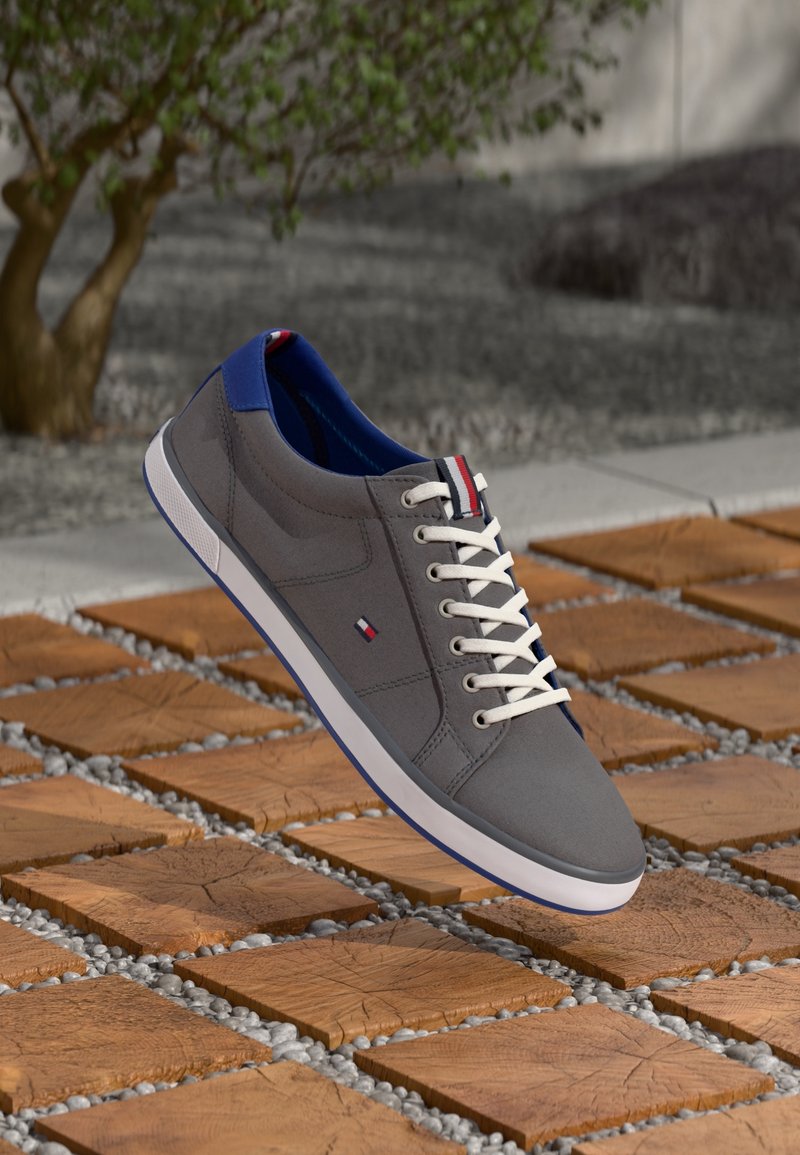 Baskets décontractées grises avec lacets blancs et doublure intérieure bleue, reposant sur une tuile en bois et une surface extérieure en galets gris.
