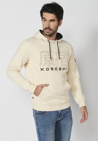 Sudadera color crema hecha de un tejido suave, con un bolsillo delantero, capucha con cordón y letras negras con textura elevada.