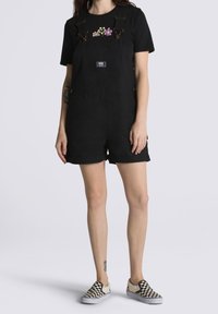 Person wearing black short overalls, black t-shirt with colorful flower designs, and black-and-white checkered slip-on shoes.