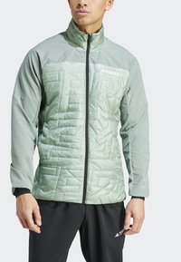 Chaqueta verde claro con acolchado en la parte delantera, cuello alto y cremallera negra. La tela es lisa con un acabado mate.