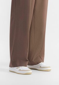 White sneakers with cream accents and textured soles paired with loose, brown, wide-leg pants, highlighting a smooth fabric finish.
