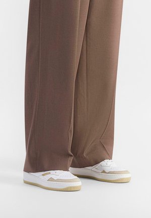White sneakers with cream accents and textured soles paired with loose, brown, wide-leg pants, highlighting a smooth fabric finish.