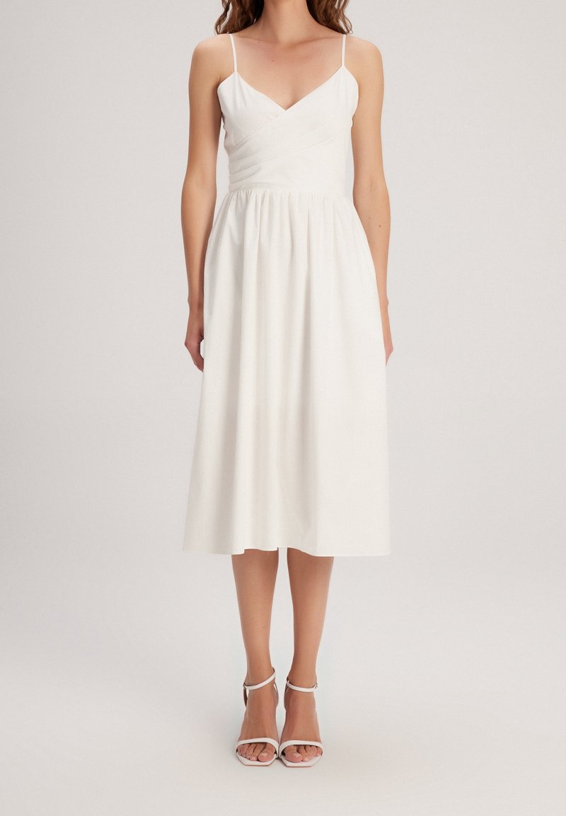 Femme portant une robe midi blanche sans manches avec fines bretelles et encolure en V, associée à des talons blancs ouverts à la cheville.