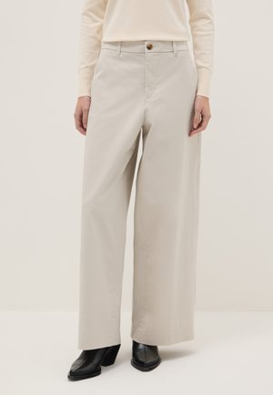 Beige wijde broek met knoop- en ritssluiting aan de voorkant, gedragen met een crèmewit shirt met lange mouwen en zwarte laarzen met hak.