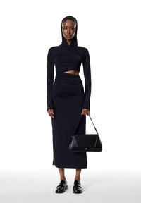 Vestido negro de manga larga con capucha, con detalles fruncidos y un corte en el abdomen. Combinado con un bolso estructurado negro y mocasines.