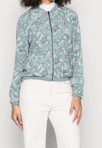 Vrouw in een lichtgroene jack met rits, bedekt met een wit bloemen- en kleurrijk vogels patroon, in combinatie met off-white broek.